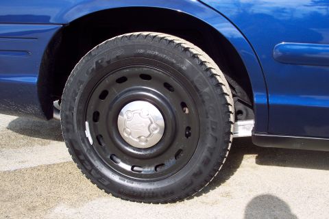 2001 Ford Crown Victoria - Close-up of a blue car's wheel and fender, showing the tire and black steel rim with a Ford logo center cap.