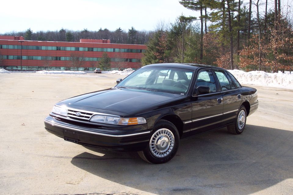 Black Ford Crown Victoria sedan parked in a lot with a brick building in the background