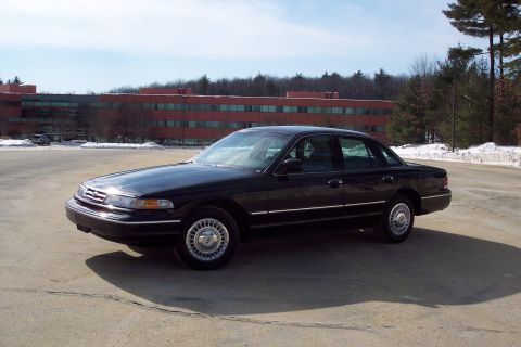 2001 Ford Crown Victoria - A black Ford Crown Victoria is parked in a lot with a large brick building in the background.
