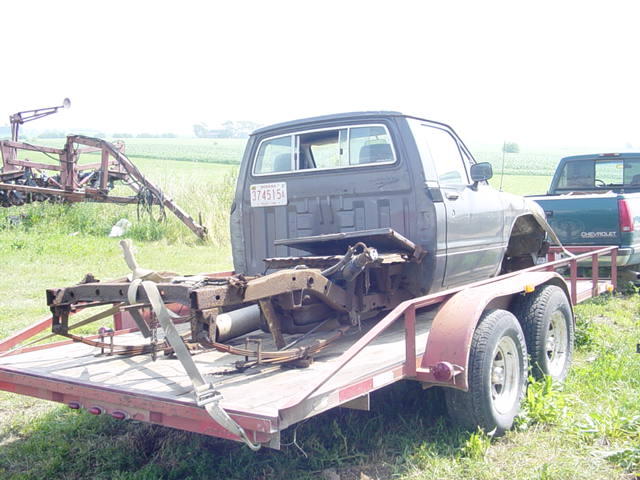 Truck chassis on a trailer, with a green Chevrolet pickup truck behind it