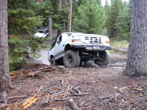 1991 Ford Bronco - A white Ford Bronco navigates a muddy trail in a forest, with another vehicle visible in the background.