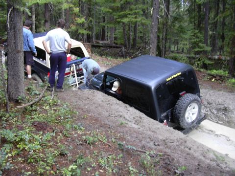 1991 Ford Bronco - Two off-road vehicles are stuck in the mud on a forest trail, with people attempting to assist.