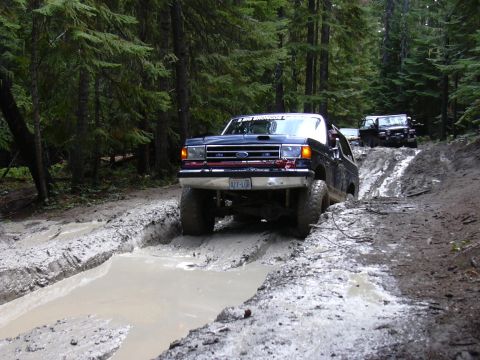 1991 Ford Bronco - A dark blue Ford Bronco is driving through a muddy trail in a forest, with another black SUV following behind.