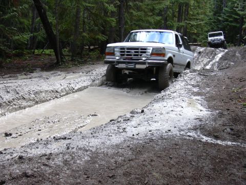 1991 Ford Bronco - A white Ford Bronco drives through a deep mud puddle on a dirt trail surrounded by trees. Another white SUV is visible further up the muddy…