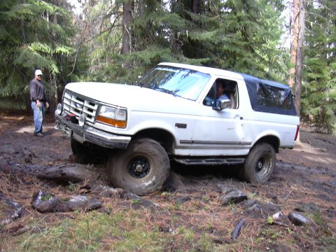 1991 Ford Bronco - A white Ford Bronco is stuck in thick mud on a wooded trail, with its front tire spinning. A man wearing a white cap and jeans stands nearb…