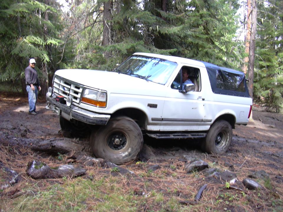 White Ford Bronco stuck in mud on a wooded trail, front tire spinning.