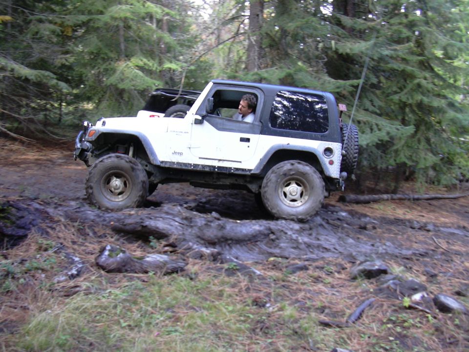 White Jeep Wrangler driving off-road on a muddy trail with a fallen log.