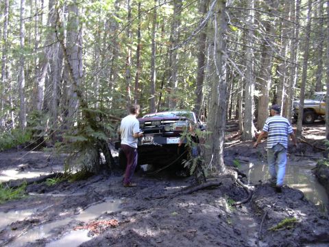 1991 Ford Bronco - Two men stand in a muddy forest clearing next to a maroon SUV that is stuck in the mud. Another SUV is visible in the background.