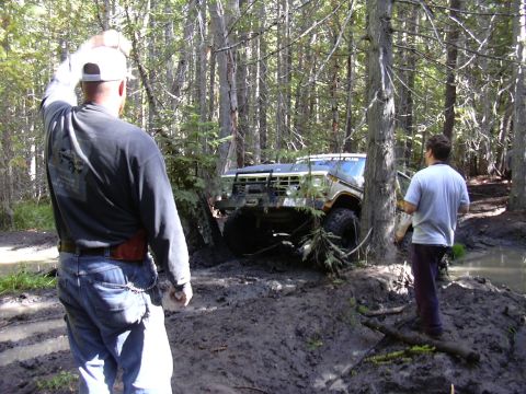 1991 Ford Bronco - Two men stand in a muddy forest clearing next to a stuck 1991 Ford Bronco. The Bronco is angled into the trees with its front end partially…
