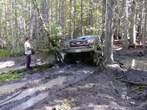 1991 Ford Bronco - A blue Ford Bronco is stuck in deep mud on a wooded trail. A person stands next to the vehicle, appearing to assess the situation.