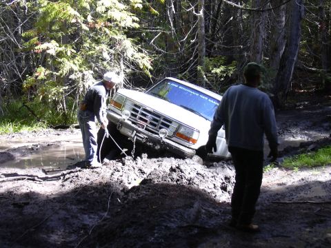 1991 Ford Bronco - Two men are attempting to pull a white Ford Bronco out of a mud pit in a wooded area. The Bronco is stuck at an angle, with its front end d…