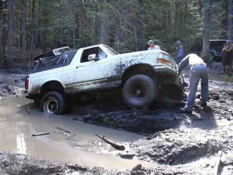 1991 Ford Bronco - A white Ford Bronco is stuck in deep mud in a wooded area. Several people are around the vehicle, appearing to be trying to free it.