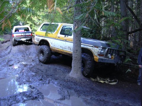 1991 Ford Bronco - Two off-road vehicles are deeply stuck in mud on a wooded trail, one being pulled by the other.