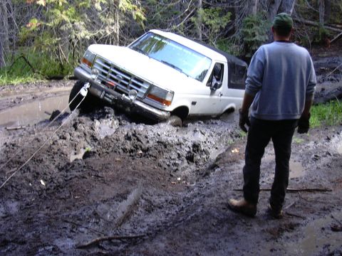 1991 Ford Bronco - A white 1991 Ford Bronco is deeply stuck in mud on a trail, with a person observing the situation.