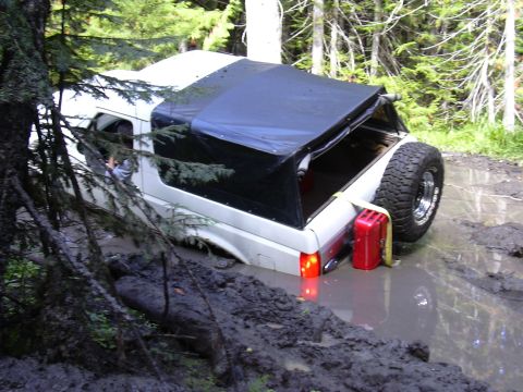 1991 Ford Bronco - A white 1991 Ford Bronco with a soft top is stuck in a muddy off-road trail.