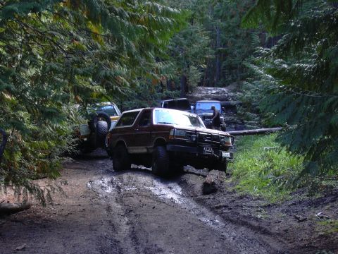 1991 Ford Bronco - A dark red Ford Bronco is navigating a muddy, wooded trail, with another SUV visible behind it. The Bronco is angled towards the viewer, sh…