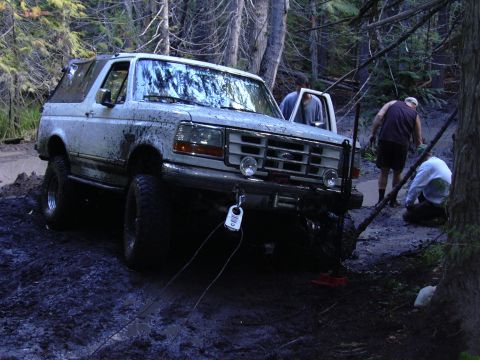 1991 Ford Bronco - A white, mud-covered Ford Bronco is stuck in a muddy forest clearing, with two people working near the front of the vehicle and another per…