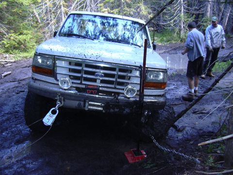 1991 Ford Bronco - A white Ford Bronco is stuck in deep mud on a wooded trail, with two men standing nearby. A winch cable is attached to the front bumper and…