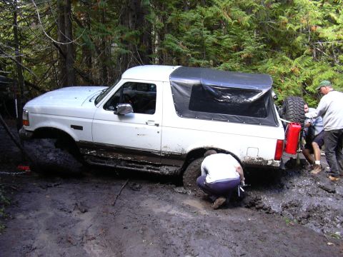 1991 Ford Bronco - A white Ford Bronco is stuck in deep mud in a forest, with several people attempting to free it. One person is kneeling by the front tire,…