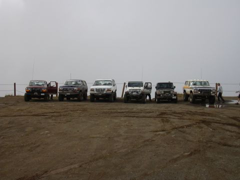 1991 Ford Bronco - A line of seven off-road vehicles, including Ford Broncos and Jeeps, are parked on a dirt lot under a hazy sky. The vehicles are positioned…
