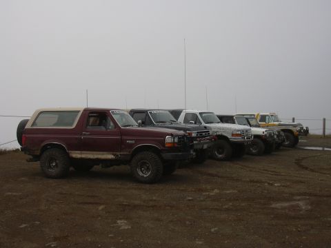 1991 Ford Bronco - A line of five off-road modified trucks and SUVs are parked on a dirt lot under a foggy sky. The closest vehicle is a maroon Ford Bronco wi…