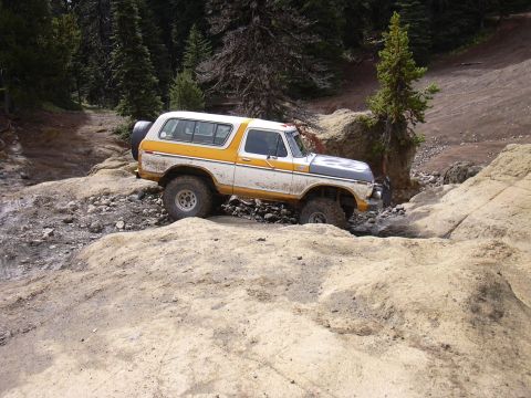 1991 Ford Bronco - A white and yellow Ford Bronco is navigating a rocky, off-road trail with dense trees in the background. The vehicle is positioned at an an…