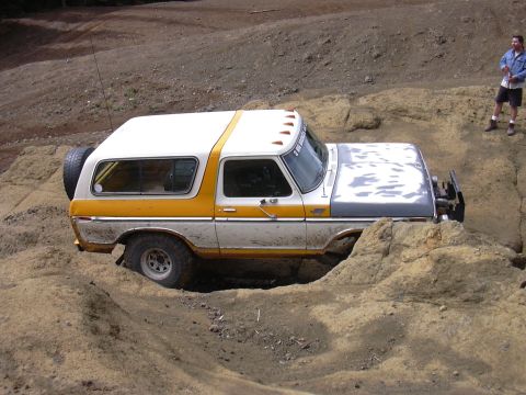1991 Ford Bronco - A two-tone yellow and white SUV is navigating a rocky, dirt terrain with its front wheel partially submerged in mud. A person in a denim ja…