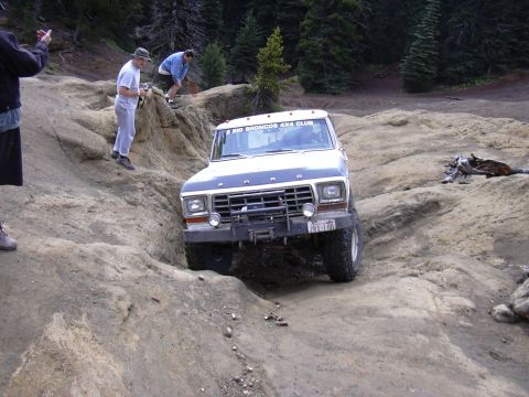 1991 Ford Bronco - A lifted, dark blue and white Ford Bronco is navigating a rocky, off-road trail with three people standing nearby. The Bronco has a black b…