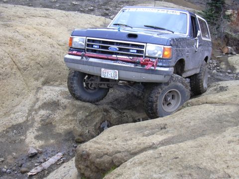 1991 Ford Bronco - A dark blue Ford Bronco is navigating a rocky, off-road trail, with its front tire significantly higher than the rear tire. A red Hi-Lift j…