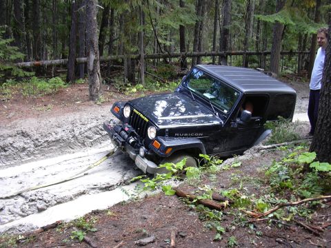 1991 Ford Bronco - A black Jeep Wrangler Rubicon is stuck in a muddy trail in a forest, with a person standing nearby.