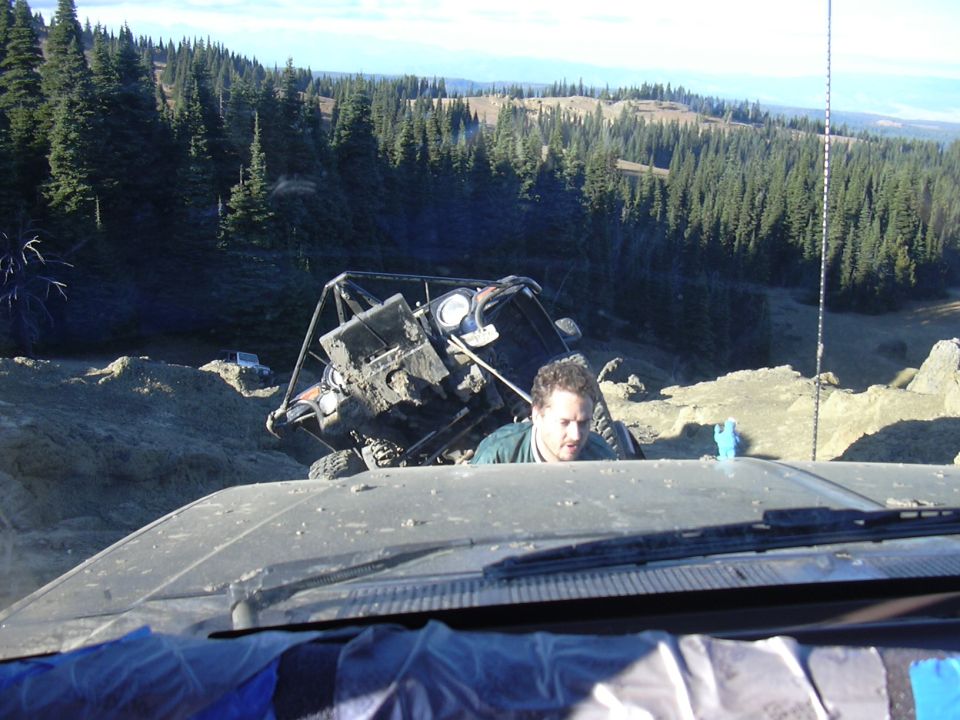 Mud-covered off-road vehicle tipped on its side on a dirt slope with a man looking out from a vehicle in the foreground