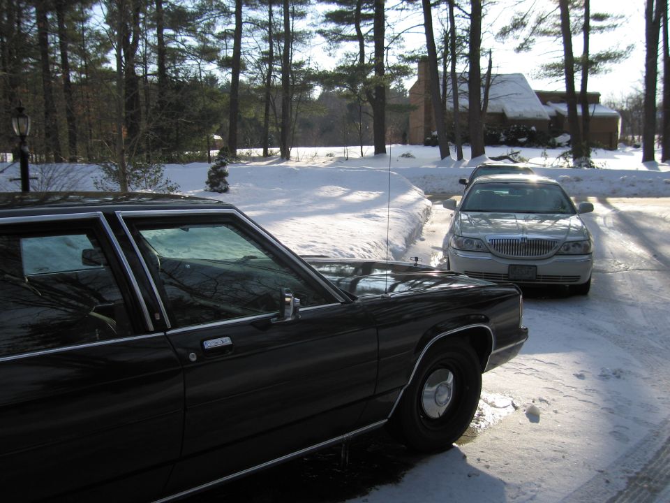 Two sedans parked in a snowy driveway with trees and a house in the background