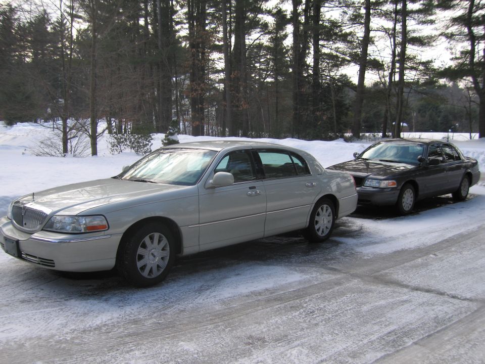 Two sedans parked on a snow-covered driveway. The front car is silver, the rear car is dark