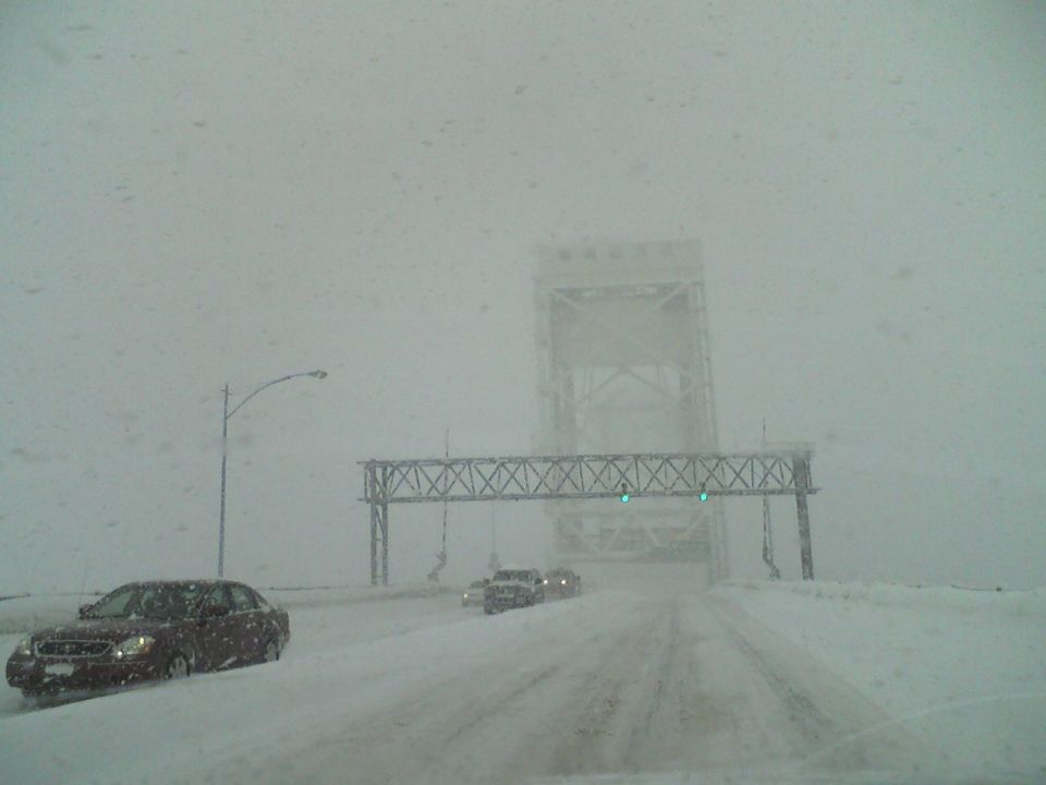 Dark sedan driving on a snow-covered road with a bridge in the background