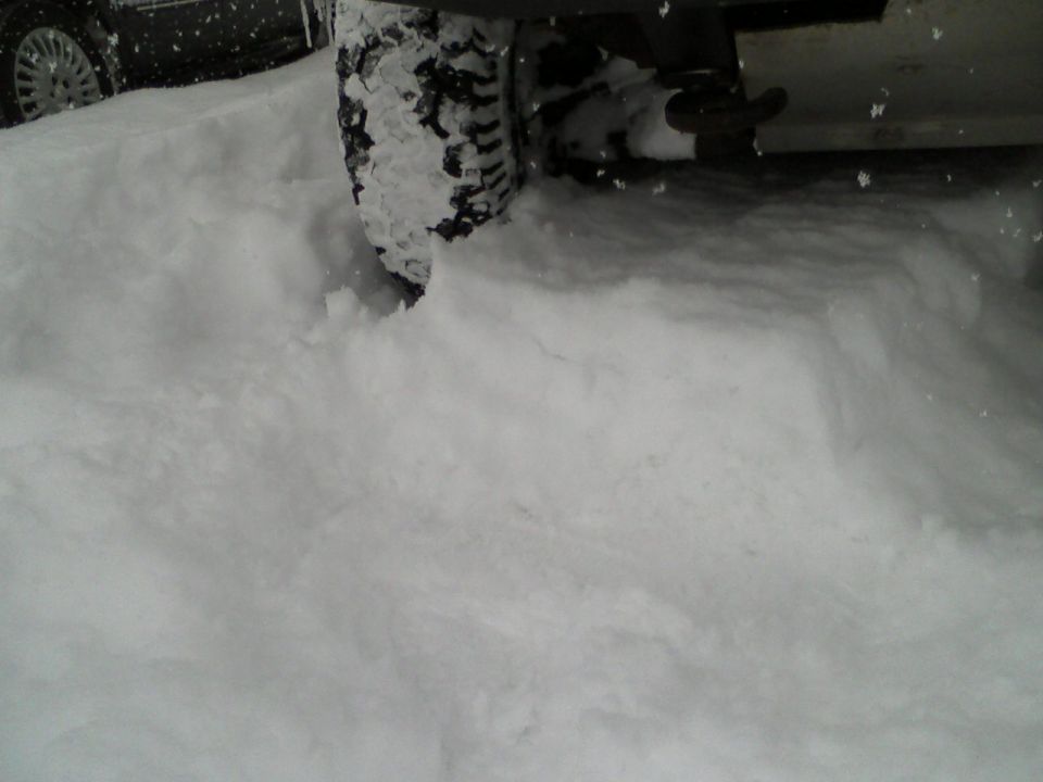 Vehicle tire partially buried in snow, another vehicle's wheel visible to the left