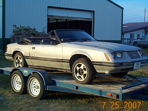 1984 Ford Mustang - A silver convertible is parked on a trailer in front of a large building.