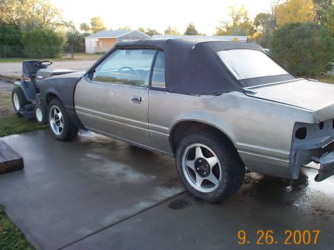 1984 Ford Mustang - A silver 1984 Ford Mustang convertible is parked outside with a lawn tractor attached to its front.