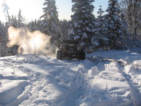 1990 Ford F-250 - An off-road vehicle navigates a snowy path through a winter forest, with steam or exhaust billowing from its front.