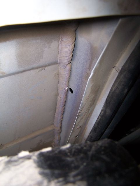 1990 Ford Bronco - Close-up view of a welded seam on the undercarriage of a vehicle, showing a small hole and a black hose nearby.