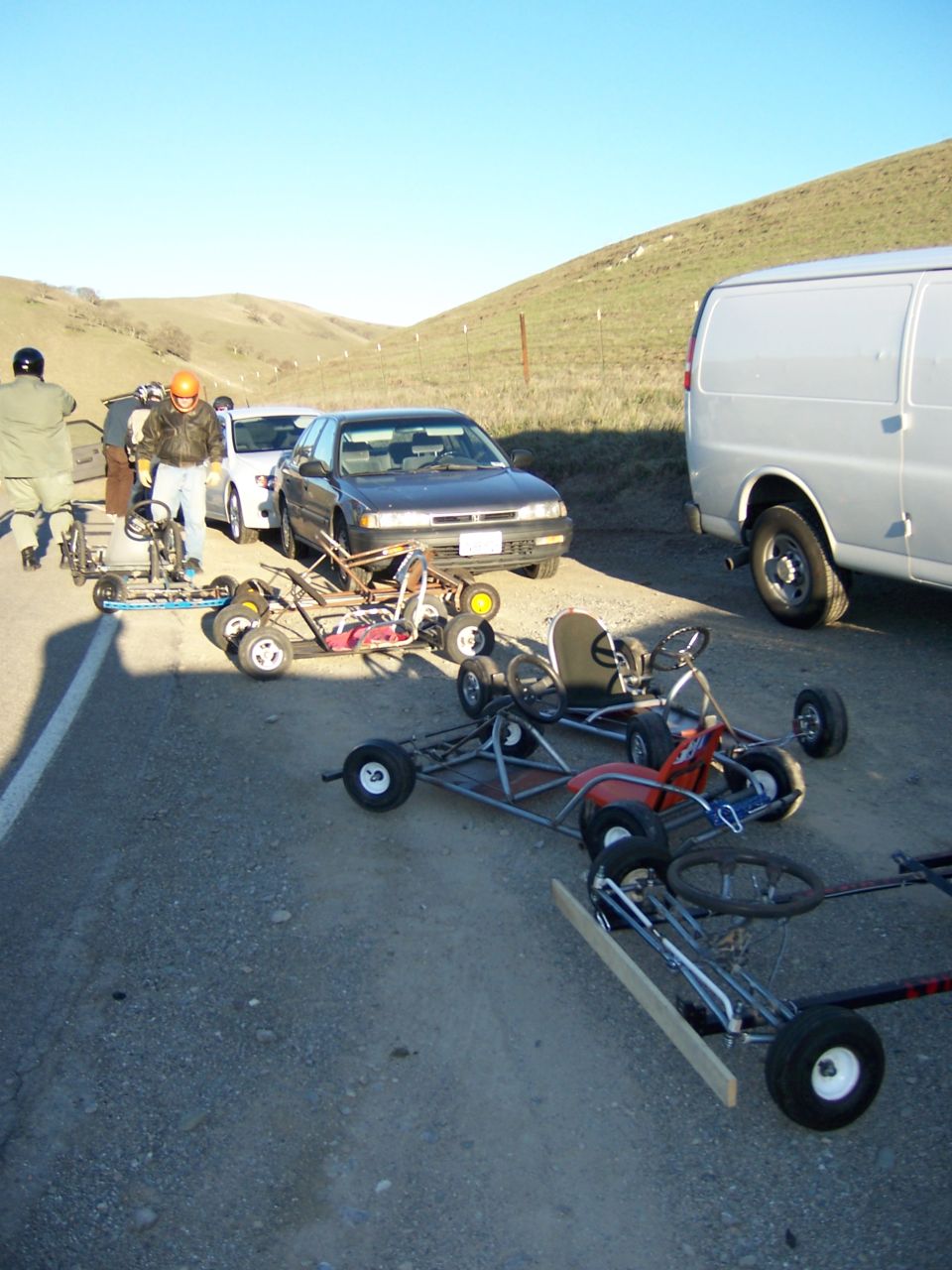 Several go-karts are lined up on a dirt road next to a sedan and a van. People in helmets are standing around