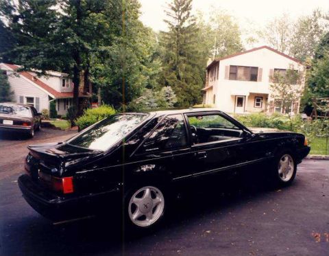 1988 Ford Mustang - A black coupe is parked in a driveway with a house and trees in the background. Another car is visible to the left.