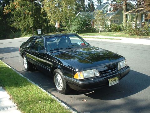 1988 Ford Mustang - A black Ford Mustang hatchback is parked curbside on a sunny day in a suburban neighborhood.