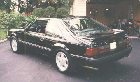 1988 Ford Mustang - A black coupe is parked outside a garage, showing its side and rear profile.