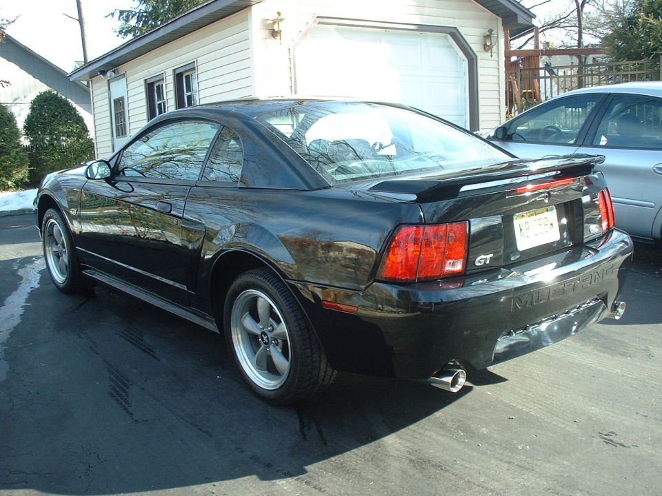 Black 2002 Ford Mustang GT coupe parked in driveway, rear view. Another car is visible to the right
