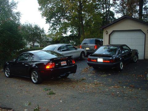 2002 Ford Mustang - Multiple vehicles including a 2002 Ford Mustang are parked in a driveway near a garage.