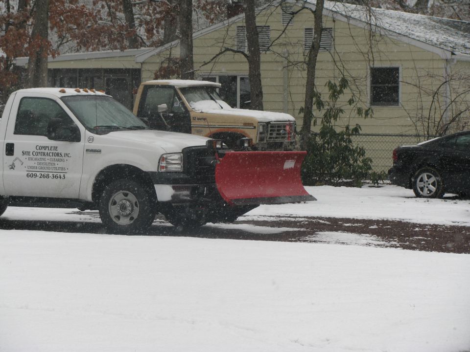 White pickup truck with red snow plow in front, another truck behind