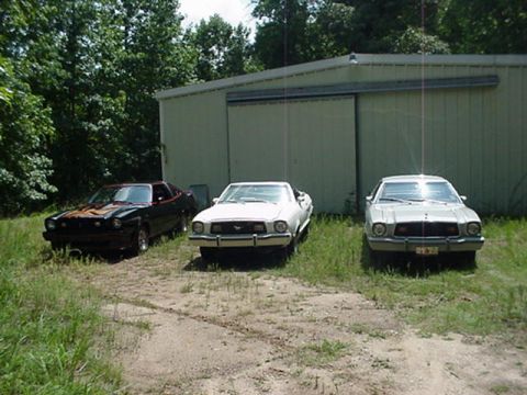 1977 Ford Mustang II - Three cars are parked in front of a large metal building. The cars are a black Mustang II, a white Mustang II, and a silver Mustang II.