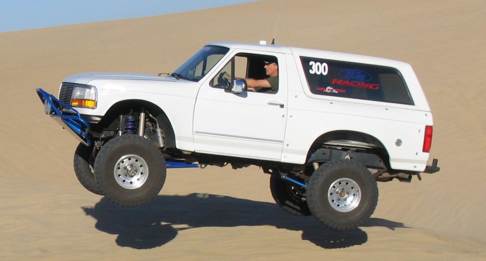 White Ford Bronco with custom blue bumper and suspension, airborne over sand dunes. It has a '300 Ford Racing' sticker on the rear window