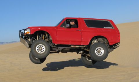 1984 Ford Bronco - Red Ford Bronco airborne over a sand dune with a driver visible.