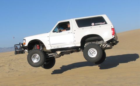 1984 Ford Bronco - This lifted SUV is airborne while traversing a sandy terrain, with another off-road vehicle visible in the distance.